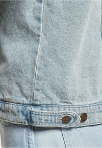 Chaqueta de denim azul claro por la parte de atrás con pestañas ajustables en la cintura aseguradas por botones metálicos grabados con "Est. 1985" y el nombre de la marca.