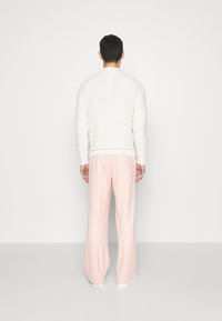 Pull en tricot blanc avec des motifs texturés, associé à un pantalon large rose clair, vu de dos, sur fond neutre.