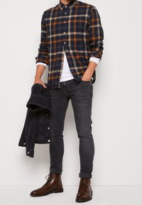 Camicia a quadri in blu navy, arancione e beige; abbottonata, a maniche lunghe. Abbinata a jeans skinny grigio scuro e stivali marroni con lacci. Giacca di jeans tenuta in mano.