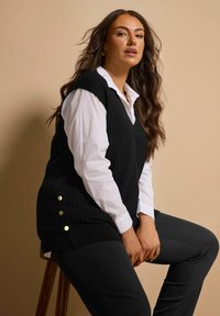 Zwarte geribbelde gebreide vest met een V-hals en gouden knopenaccenten aan de zijkanten, gedragen over een witte blouse met kraag en gecombineerd met zwarte broek.