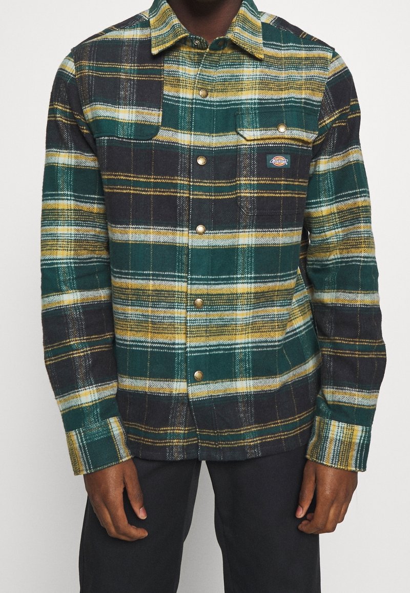 Homme portant une chemise à manches longues à carreaux verts, jaunes, noirs et blancs avec poches poitrine et pantalon sombre.