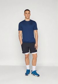 Marineblå sportst-shirt med korte ærmer, kombineret med sorte shorts med hvid kant. Modellen har blå sportssko og hvide sokker på.