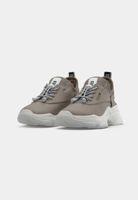 Un paio di sneaker in rete color taupe con suole bianche spesse e lacci elastici grigi senza nodo, posizionate su uno sfondo bianco semplice.