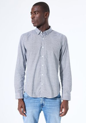 Mann trägt hellgraues Hemd mit Muster und blaue Jeans, schaut nach links vor weißem Hintergrund.