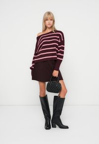 Un pull en tricot surdimensionné aux rayures bordeaux et roses, une jupe sombre, des bottes noires jusqu'aux genoux et un petit sac à main noir texturé.
