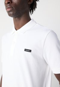 Das weiße Baumwoll-Polohemd verfügt über einen klassischen Kragen, drei Knöpfe und ein schwarzes rechteckiges Calvin Klein Logo-Patch auf der linken Brust.