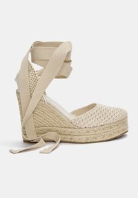 Beige kilklack-espadrilles med vävt tyg ovanpå, texturerad jute mellanläggssula och tygremmar vid halvkalven för säker fastsättning.