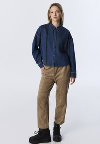 Denim overhemd met knopen en contrasterende stiksels, enkele borstzak; gedragen met tan suède broek en zwarte gestippelde plateauschoenen.