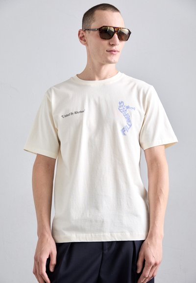T-shirt en coton couleur crème à manches courtes. L'avant présente le texte "United By Rhythm" et un élément graphique bleu.