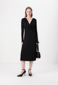Black long-sleeve dress with a deep V-neck and twist detail at the bodice, midi length, worn with pointed-toe heels and a small handbag.