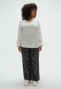 Λευκό μπλουζάκι με μακριά μανίκια και V-λαιμόκοψη, συνδυασμένο με φαρδιά μπλε σκούρα παντελόνια με γεωμετρικά λευκά σχέδια και λευκά αθλητικά παπούτσια.