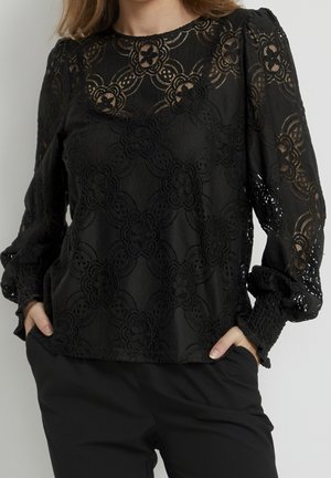 Black long-sleeve lace blouse with floral patterns, puffed sleeves, and smocked cuffs worn with black pants, hands in pockets.