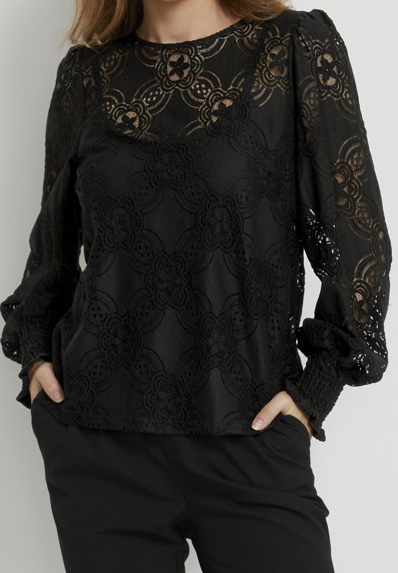 Blouse noire en dentelle manches longues avec motifs floraux, manches ballon et poignets smockés portée avec un pantalon noir, mains dans les poches.