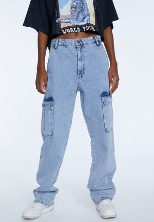 Person iført lyseblå, højtaljede denim cargobukser med klaplommer og hvide sneakers, kombineret med en cropped sort grafisk T-shirt.
