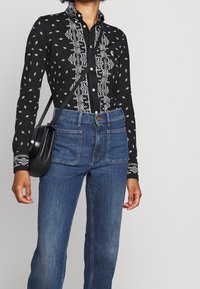 Svart långärmad tröja med vita geometriska mönster och broderade detaljer, i kombination med högmidjade blå denimjeans. Svart axelremsväska.