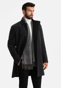 Abrigo de lana negra con diseño de doble botonadura, cuello alto y botones grandes. Bufanda gris a cuadros con flecos llevada por debajo.