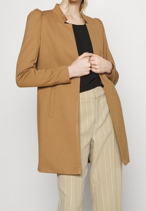 Personne portant un blazer couleur camel avec des épaules bouffantes, un top noir et un pantalon beige à fines rayures, tenant les revers du blazer.
