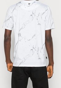 Camiseta blanca con patrón de mármol en gris y negro. Mangas cortas, cuello redondo y pequeño detalle del logo en la parte trasera del cuello. Tela ligera.
