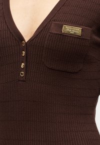 Καφέ τοπ από ριπ υφανσία με βαθύ V-λαιμόκοψη, τέσσερα κουμπιά σε χρυσό τόνο και μια μικρή τσέπη με διακοσμητικό χρυσό ταμπελάκι.