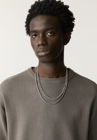 Grauer Pullover mit rundem Ausschnitt und zwei silbernen Halsketten. Die Textur ist weich, mit Rippendetails an den Ärmelbündchen und am Kragen.