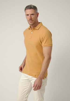 Hombre con cabello corto y gris que lleva una camiseta polo amarilla mostaza y pantalones blancos, de pie frente a un fondo claro y liso.