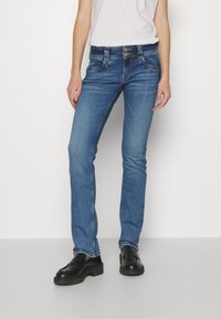 Jeans de mezclilla azul oscura con corte ajustado, cintura a media altura, estilo de cinco bolsillos y costuras visibles. Combinados con zapatos negros.