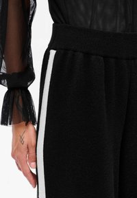 Zwarte joggerbroek met een zachte textuur, elastische tailleband en een witte zijstreep. Een hand met een tatoeage is op de broek geplaatst.