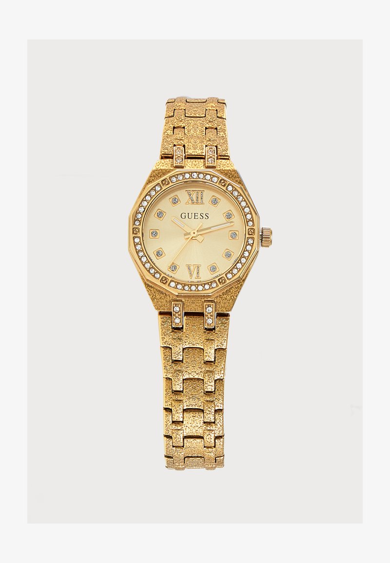 Χρυσού χρώματος ρολόι χειρός Guess με σημεία ώρας από κρύσταλλα, ρωμαϊκούς αριθμούς στις 12 και 6, και υφασμένη μεταλλική σύνδεση.