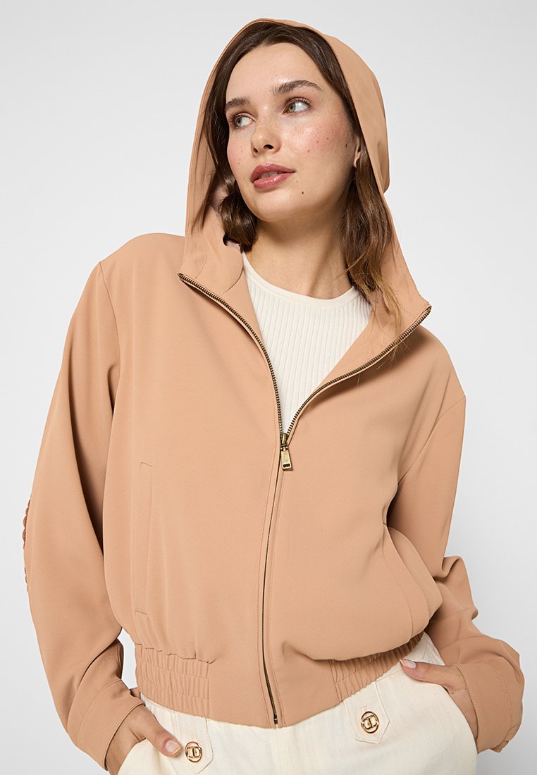 Sudadera con cremallera de color marrón claro, hecha de un material suave, con capucha, cinturilla elástica acanalada y bolsillos laterales. Textura suave, diseño minimalista.