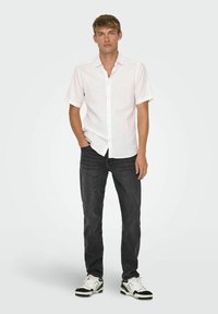 Camisa blanca de manga corta y botón con un ajuste relajado, combinada con jeans oscuros y zapatillas en blanco y negro. Diseño simple sin patrones.