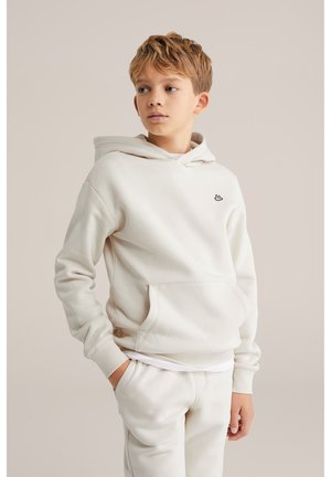 Fiú krémszínű kapucnis pulóverben és hozzá illő nadrágban, egyik kezét zsebében tartva, oldalra nézve, egyszínű háttér előtt áll.