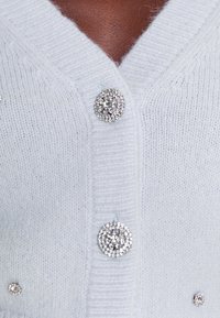 Gros plan sur un cardigan blanc en tricot avec des boutons ronds en strass et des strass décoratifs sur un tissu doux.