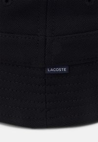 Svart vevd stofflue med Lacoste-merke, teksturert overflate og subtile sømmer. Rund form med en strukturert passform.