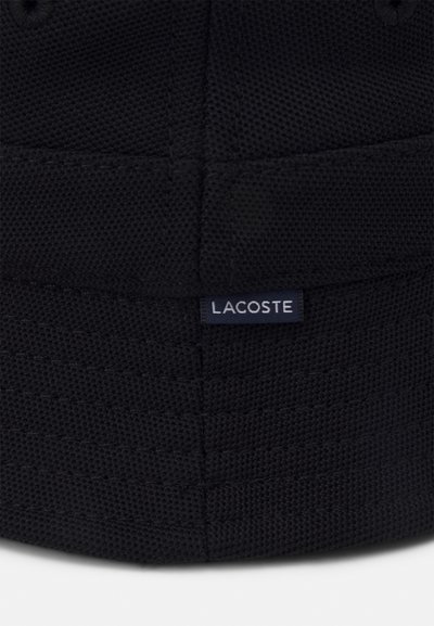 Casquette en tissu tissé noir avec une étiquette Lacoste, surface texturée et détails de couture subtils. Forme ronde avec un ajustement structuré.