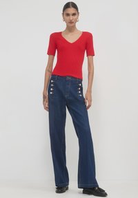 Frau mit zurückgebundenem Haar, die ein rotes V-Ausschnitt-Shirt mit kurzen Ärmeln, hochgeschnittene, weite, dunkelblaue Jeans mit Knöpfen und schwarze Slipper trägt.