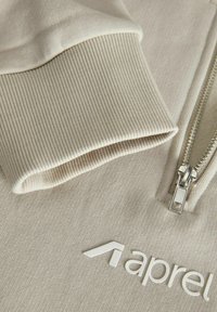 Artimo vaizdas: smėlio spalvos dryžuoto audinio rankogalis ir sidabrinė užtrauktukas ant šviesiai smėlio spalvos drabužio su daliniu baltu „apparel“ logotipu.