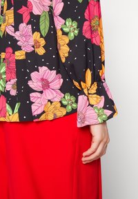 Blusa di tessuto nera con stampa floreale colorata in rosa, arancio e verde. Caratterizzata da un polsino arricciato e una vestibilità ampia. Abbinata a una gonna rossa.