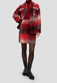 Femme portant une jupe à carreaux rouges et noirs, une veste à carreaux oversize assortie, un col roulé bordeaux, des collants noirs et des bottines noires.