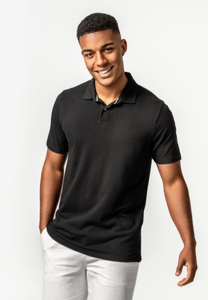 Lächelnder junger Mann mit kurzen lockigen Haaren, der ein schwarzes Poloshirt und weiße Hose trägt und vor einem einfarbigen hellen Hintergrund steht.