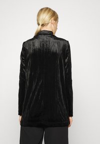 Černý sametový blazer s měkkým, texturovaným povrchem. Má přizpůsobený střih, dlouhé rukávy a klopový límec.