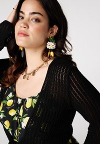 Een vrouw met lang, donker haar en een peinzende uitdrukking draagt een zwarte cardigan over een top met citroenenpatroon, aangevuld met bloemenoorbellen.