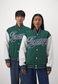 Jaquetas varsity verde e branca com um proeminente logotipo "Tommy". Apresenta punhos e gola canelados, fechos de botão seguros e um corte descontraído.