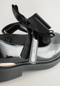 Chaussure en cuir verni noir avec semelle plate, dotée d'un détail en nœud sur la sangle et d'un accent en ruban texturé. Surface lisse, design minimaliste.
