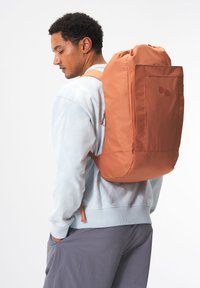 Oranger Rucksack mit einem Zugverschluss und einer großen Fronttasche. Aus weichem Stoff gefertigt, hat er zwei runde Akzente und verstellbare Träger.