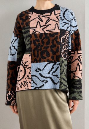Femme portant un pull patchwork multicolore avec des motifs d’étoile, de soleil, d’oiseau et d’animaux abstraits, associé à une jupe vert olive.