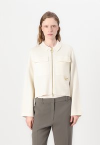 Κρεμ χρώματος κοντό μπουφάν με γιακά, διαθέτει μπροστινό φερμουάρ, δύο τσέπες στο στήθος και χρυσές λεπτομέρειες. Λείος υφασμάτινος ιστός.