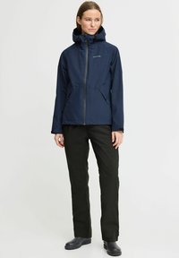Marineblauer wasserdichter Anorak mit Reißverschluss, verstellbarem Kapuzen und zwei Seitentaschen, kombiniert mit schwarzen Hosen und schwarzem Schuhwerk.