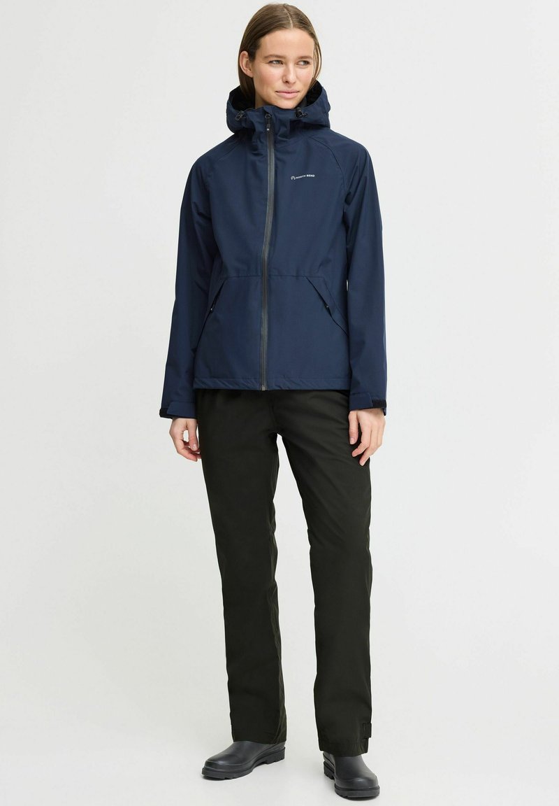 Marineblauer wasserdichter Anorak mit Reißverschluss, verstellbarem Kapuzen und zwei Seitentaschen, kombiniert mit schwarzen Hosen und schwarzem Schuhwerk.