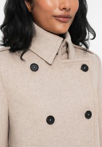Cappotto in misto lana beige con design doppiopetto, grandi bottoni neri, colletto alto e una finitura morbida e testurizzata.