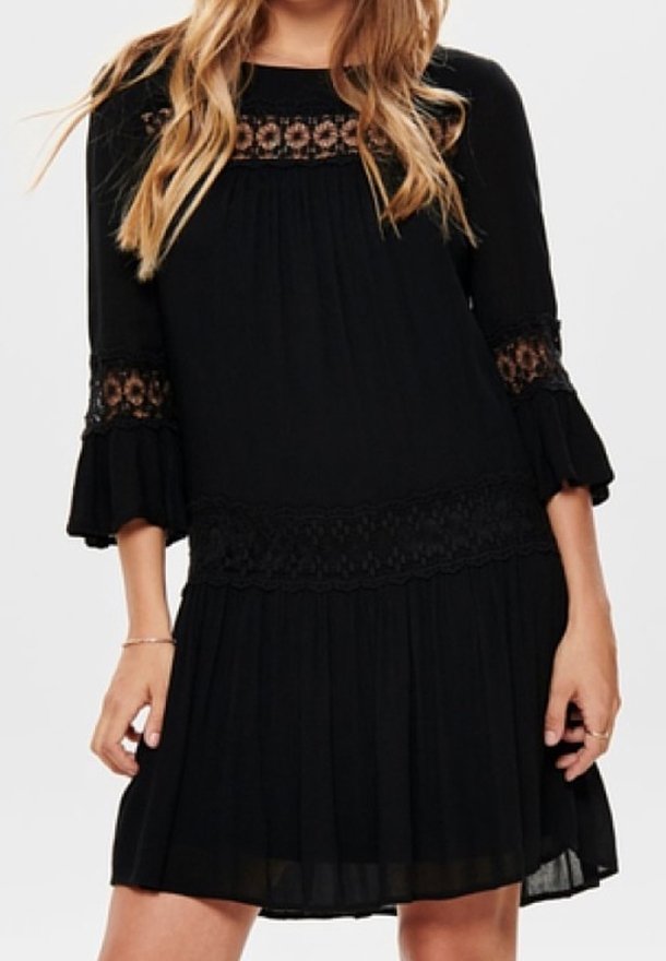 Femme portant une robe noire avec des détails en dentelle sur la poitrine, les manches et la taille, avec des poignets évasés et une jupe plissée.